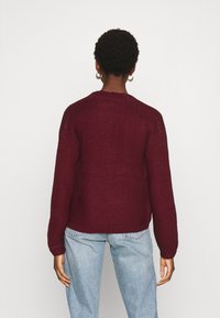 Burgundyröd stickad tröja med långa ärmar, rund halsringning och strukturerad yta. Bärs tillsammans med ljusblå jeans, sedd från baksidan.