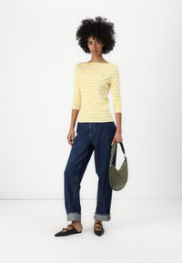 Gelb-weiß gestreiftes Langarmhemd kombiniert mit blauen weit geschnittenen Jeans. Das Model trägt eine grüne Handtasche und schwarze Ballerinas.