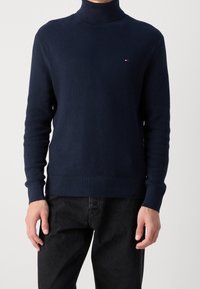 Homme portant un pull à col roulé texturé bleu marine avec un petit logo rouge, blanc et bleu sur la poitrine et un pantalon noir.