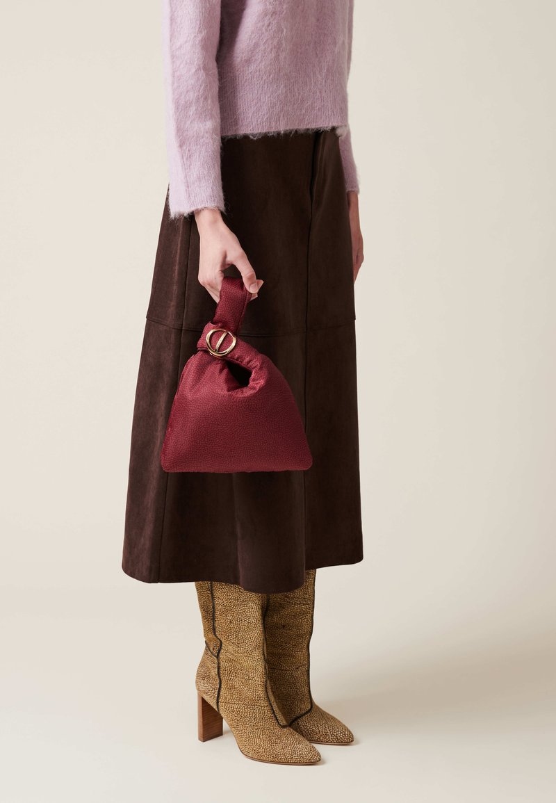 Burgundy textured handbag with a knot-style handle featuring a gold accent. Paired with a brown suede skirt and patterned heeled boots.