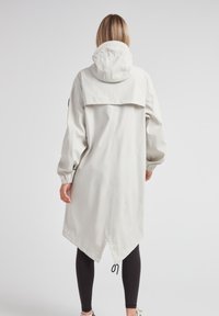 Cappotto lungo impermeabile grigio chiaro con cappuccio, polsini elastici e un orlo angolato unico. Presenta una fessura posteriore per maggiore libertà di movimento.