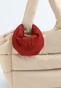 Sac fourre-tout matelassé beige avec une petite pochette zippée rouge attachée à une poignée, sur un fond uni.