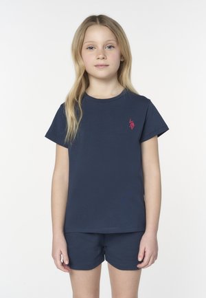 Ragazza giovane con lunghi capelli biondi che indossa una maglietta blu navy e pantaloncini abbinati, in piedi davanti a uno sfondo bianco semplice.