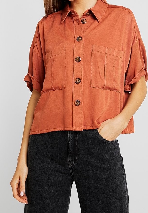 Lyhythihainen, ruosteenvärinen paita, joka on valmistettu pehmeästä kankaasta. Paidassa on napillinen etuosa, terävä kaulus ja kaksi rintataskua.