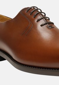 Chaussure de ville en cuir brun avec une finition lisse, ornée de détails de coutures droites et de lacets marron sur un bout rond et un talon bas.