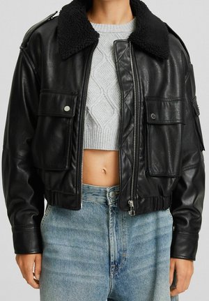 Person wearing a black leather jacket with a shearling collar over a gray cropped cable-knit sweater and high-waisted blue jeans.