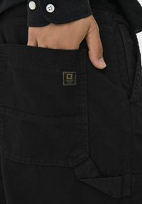 Zwarte cargobroek van stof met een gestructureerde afwerking, voorzien van meerdere zakken en een klein merkmine. Hand in een zijzak gestoken.