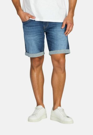 Man wearing rolled-up blue denim shorts, white sneakers, and a white shirt, standing with one hand in pocket.