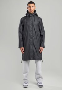 Long black waterproof coat with a hood, featuring snap buttons, two side pockets, and drawstrings at the neck and hem.
