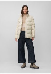 Žena oblečená v béžovom zateplenom kabáte, béžovom svetri, širokých tmavých džínoch a šedých hrubých topánkach stojí proti jednoduchému šedému pozadiu.