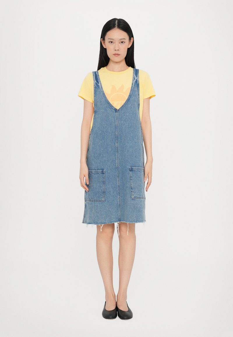 Robe en denim à bretelles avec un décolleté en V, deux grandes poches avant, un ourlet brut et sans manches, portée par-dessus un t-shirt graphique jaune.