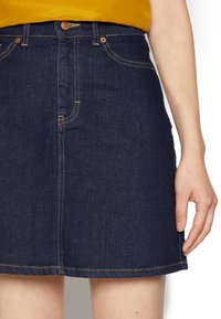 Saia de ganga, azul escuro, com uma forma ligeiramente evasé. Apresenta costuras laranja e acabamentos em botão de metal na cintura.