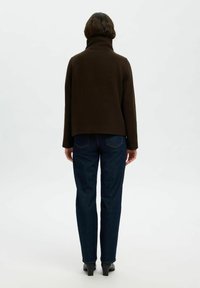 Pullover marrone con colletto alto e vestibilità rilassata, abbinato a jeans blu scuro. Il materiale sembra spesso e strutturato, evidenziando la semplicità nel design.