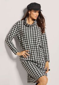 Kleid mit Hahnentrittmuster in Schwarz und Weiß, mit einem Schalkragen, langen Ärmeln, lockerer Passform und seitlichem Schlitz für zusätzlichen Komfort. Mit einer schwarzen Mütze accessoriert.