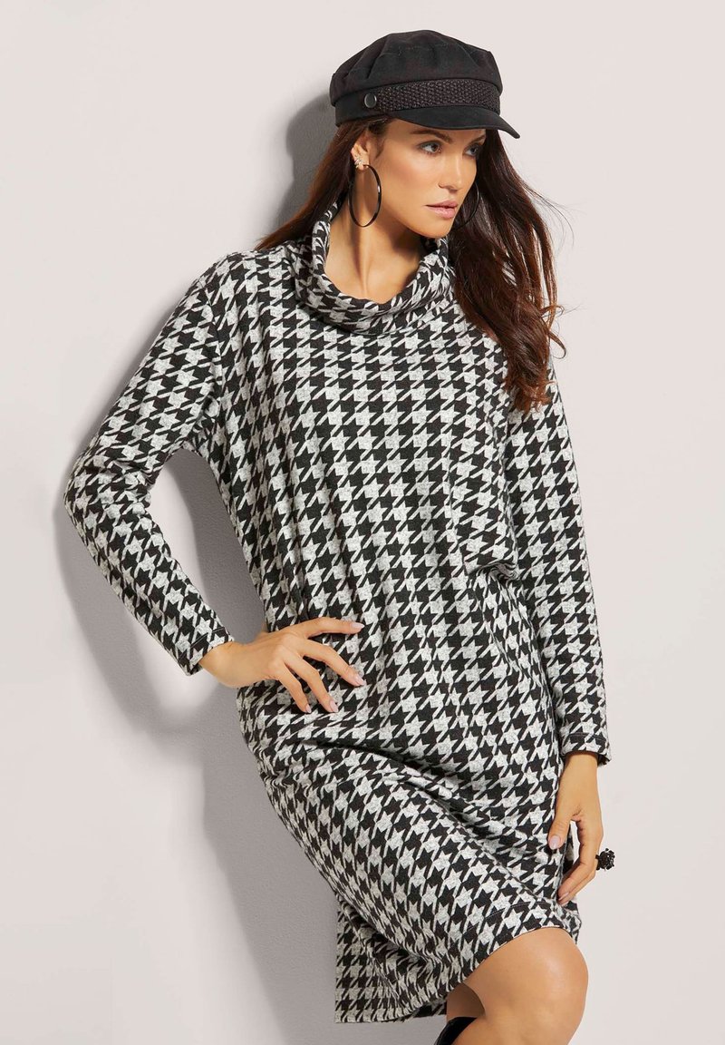 Kleid mit Hahnentrittmuster in Schwarz und Weiß, mit einem Schalkragen, langen Ärmeln, lockerer Passform und seitlichem Schlitz für zusätzlichen Komfort. Mit einer schwarzen Mütze accessoriert.