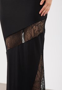 Μαύρο φόρεμα με εφαρμοστή γραμμή και ανοίγματα από δαντέλα. Διαθέτει λείο ύφασμα και λεπτές ανθικές δαντέλες κατά μήκος του ποδόγυρου.