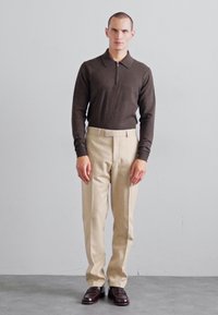 Polo marrón de manga larga con cuello de zip, combinado con pantalones beige ajustados y zapatos burdeos oscuros. Tejido texturizado, diseño estilizado.