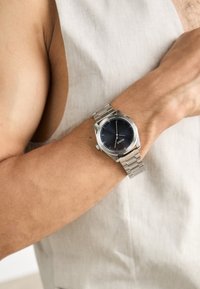 Reloj de acero inoxidable plateado con una esfera redonda negra, diseño minimalista y pulsera de eslabones, llevado en una muñeca sobre un fondo de tela clara.