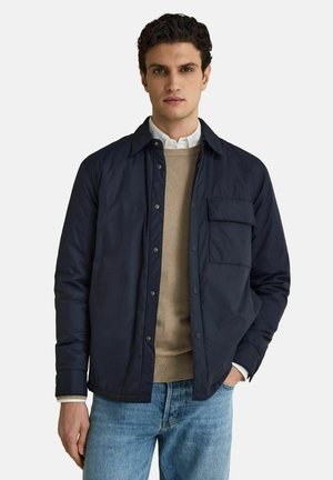 Man i marinblå vadderad jacka över beige tröja och vit skjorta, med ljusblå jeans, stående med ena handen i fickan.