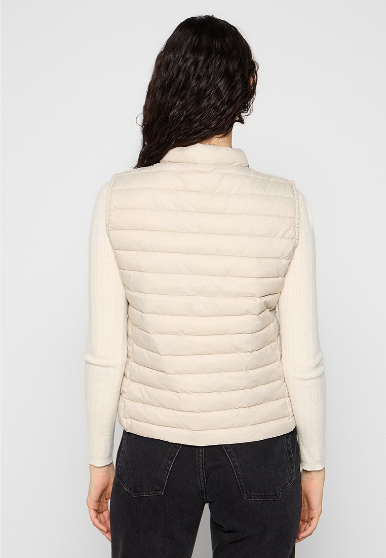Frau mit dunklen lockigen Haaren, die eine hellbeige gesteppte Weste über einem cremefarbenen langärmligen Pullover und schwarzen Jeans trägt, steht vor einem schlichten Hintergrund.