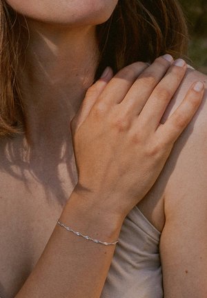 Silberner, verdrehter Armband, der an einem gebräunten Unterarm getragen wird. Der Hautton ist warm und der Hintergrund ist weichgezeichnet, was das Schmuckstück hervorhebt.