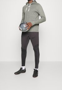 Haut à capuche long gris clair avec des cordons blancs, associé à un pantalon de sport gris foncé avec des accents texturés et des crampons de football noirs.