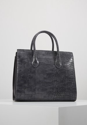 Sac à main structuré noir avec texture crocodile, deux poignées arrondies, posé sur une surface blanche devant un fond gris.