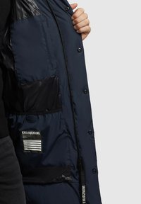 Tumedast puffer jakk, mis on läikiva viimistlusega, mitme tasku, bränditud sildiga ja esiosal tõmblukuga, millel on nööpkinnitus. Tekstureeritud kangas sisemus.