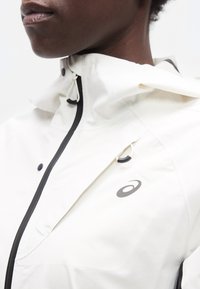 Κοντινό πλάνο ενός ατόμου που φοράει λευκό αδιάβροχο μπουφάν με μαύρο φερμουάρ, κουκούλα και ένα μικρό μαύρο λογότυπο στο στήθος.