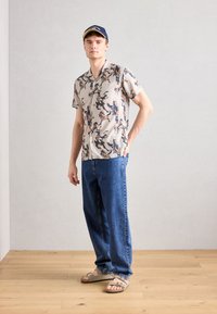 Short-sleeve beige shirt with a multi-colored abstract pattern, paired with loose blue jeans and tan sandals. Casual hat enhances the look.