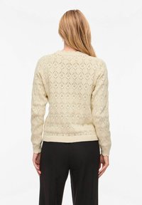 Femme aux cheveux blonds longs portant un pull crème en maille avec un motif géométrique et un pantalon noir, vue de dos sur un fond blanc.