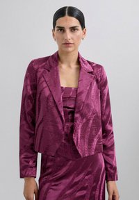 Blazer ajustado y recortado en vibrante magenta con un patrón texturizado y ondulado. Presenta solapas de muesca y mangas largas, usado sobre una blusa a juego.