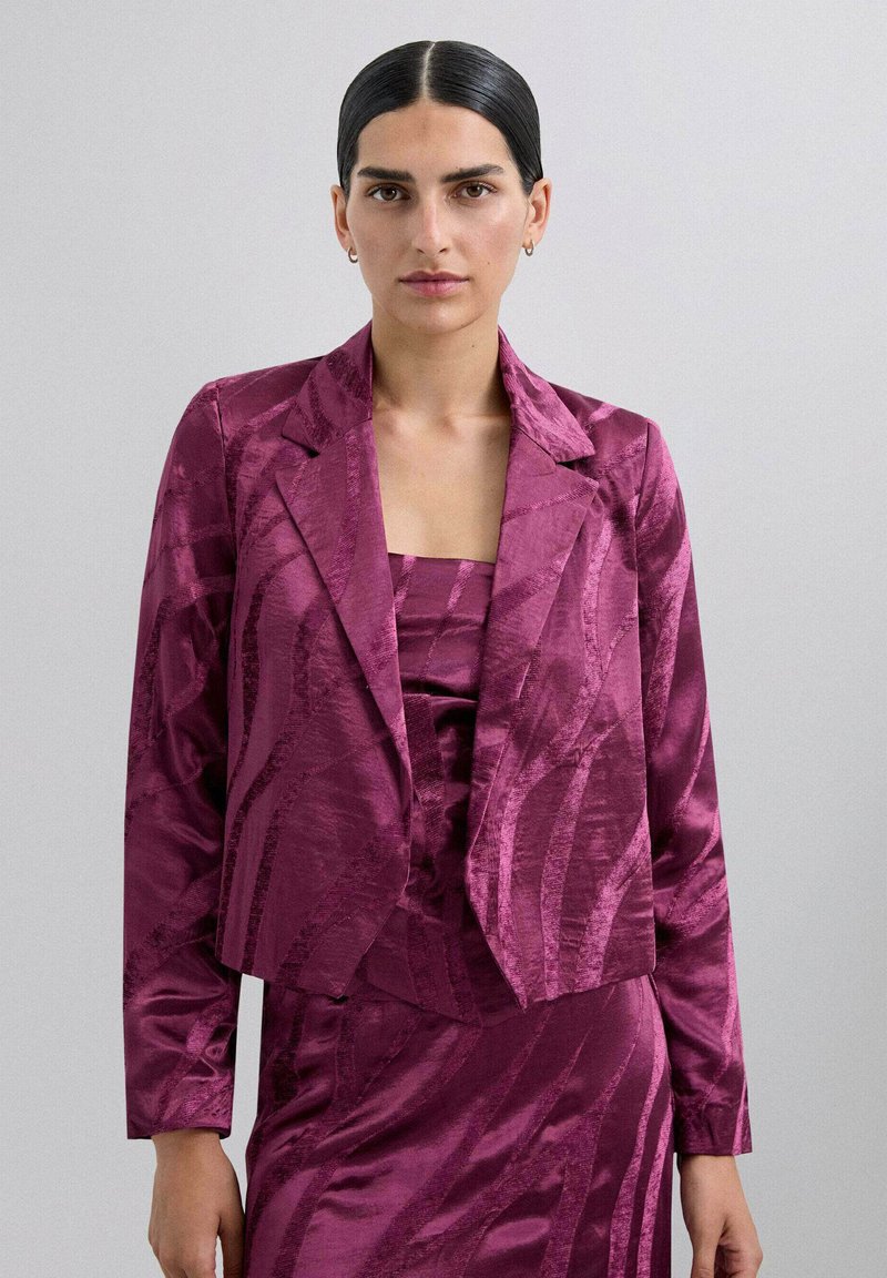 Blazer ajustado y recortado en vibrante magenta con un patrón texturizado y ondulado. Presenta solapas de muesca y mangas largas, usado sobre una blusa a juego.