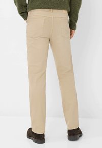 Pantaloni in cotone beige con gamba dritta, tasche posteriori visibili e cuciture a contrasto, indossati con stivali alla caviglia marrone scuro.