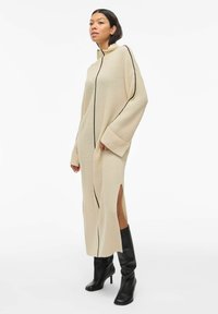 Robe en tricot beige avec un col montant, des bordures noires sur les côtés et des fentes latérales ; des manches larges et une texture structurée ajoutent de l'intérêt visuel.