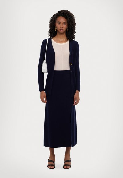 Cardigan a coste blu navy, top beige chiaro, gonna blu con spacco, sandali neri con tacco, piccola borsa bianca, tessuto testurizzato. Design minimalista.