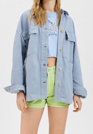 Femme portant une veste surdimensionnée bleu clair par-dessus un top graphique bleu court et un short vert vif, debout devant un fond uni.