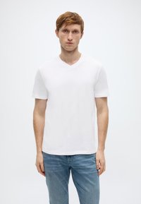 Λευκό κοντομάνικο μπλουζάκι V λαιμόκοψης από βαμβάκι, παρουσιασμένο σε μοντέλο που φοράει μπλε τζιν, απλό φόντο.