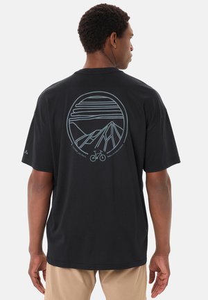 Von hinten gesehener Mann, der ein schwarzes T-Shirt mit einer Zeichnung von Berg, Fahrrad und Himmel auf dem Rücken trägt, beige Shorts.