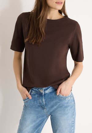 Femme portant un t-shirt à manches courtes marron foncé uni avec un jean en denim bleu clair, les mains dans les poches, debout devant un fond uni.