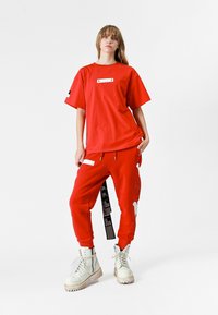 Camiseta roja de gran tamaño con etiqueta blanca, combinada con pantalones de chándal rojos con acentos gráficos y una etiqueta negra prominente. Botas de combate blancas.