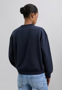 Sudadera azul marino con un corte relajado, cuello de tripa acanalado y mangas largas; la tela parece suave y texturizada; vista trasera.