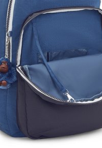 Blauwe stoffen rugzak met een donkerblauwe onderkant, voorzien van een ritsvak aan de voorkant en een harige aap sleutelhanger bevestigd aan de rits.