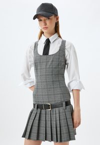 Vestido mini a cuadros grises con escote cuadrado, cinturón negro y falda con plisados. Llevado sobre una camisa blanca y combinado con una gorra gris.