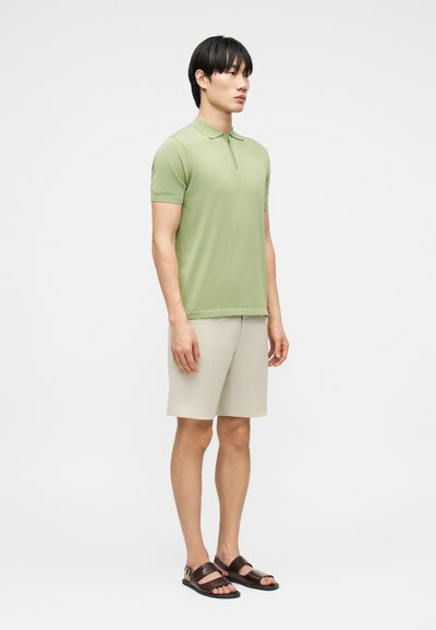 Homme debout portant un polo à manches courtes vert clair, un short beige et des sandales marron foncé sur un fond blanc uni.