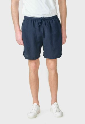 Mand iført marineblå shorts med snøre, hvid t-shirt og hvide sneakers står mod en ensfarvet baggrund.