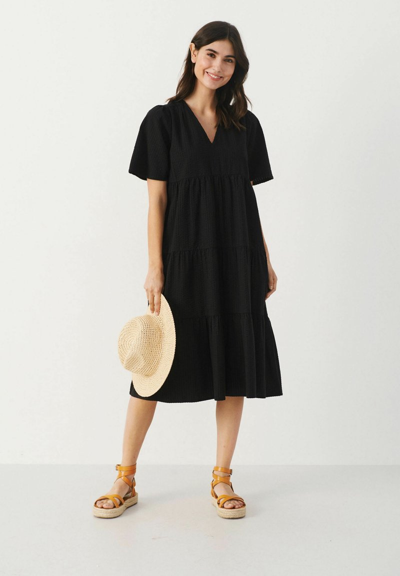 Schwarzes, gestuftes Kleid mit kurzen Ärmeln und V-Ausschnitt. Waffelstruktur und lockere Passform. Kombiniert mit sandfarbenen Riemchensandalen und einem Strohhut.