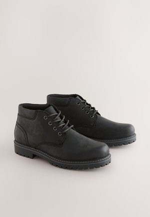 Fekete bőr bokacsizma kerek orral, hat fűzőlyukkal és vastag gumitalppal. Kontrasztos varrással és texturált felülettel rendelkezik.