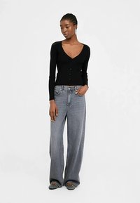 Črn kardigan z dolgimi rokavi na gumbe in z V-izrezom, kombiniran s svetlo sivimi širokimi kavbojkami in ravnimi čevlji.