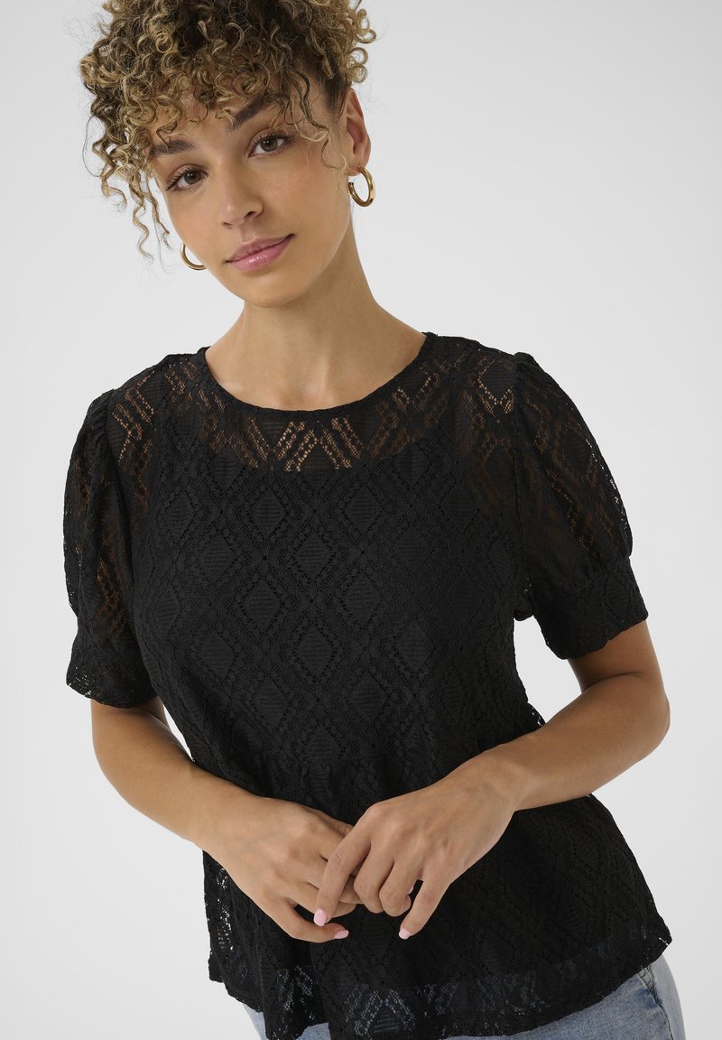 Top in pizzo nero con motivo a diamante, maniche a puff corte, scollatura rotonda e orlo smerlato. Le mani della modella sono posizionate in vita.